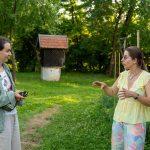 Două femei stau de vorbă pe iarbă, în fața unei case albe; una dintre ele ține un aparat foto