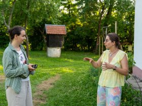 Două femei stau de vorbă pe iarbă, în fața unei case albe; una dintre ele ține un aparat foto