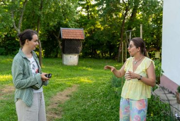 Două femei stau de vorbă pe iarbă, în fața unei case albe; una dintre ele ține un aparat foto