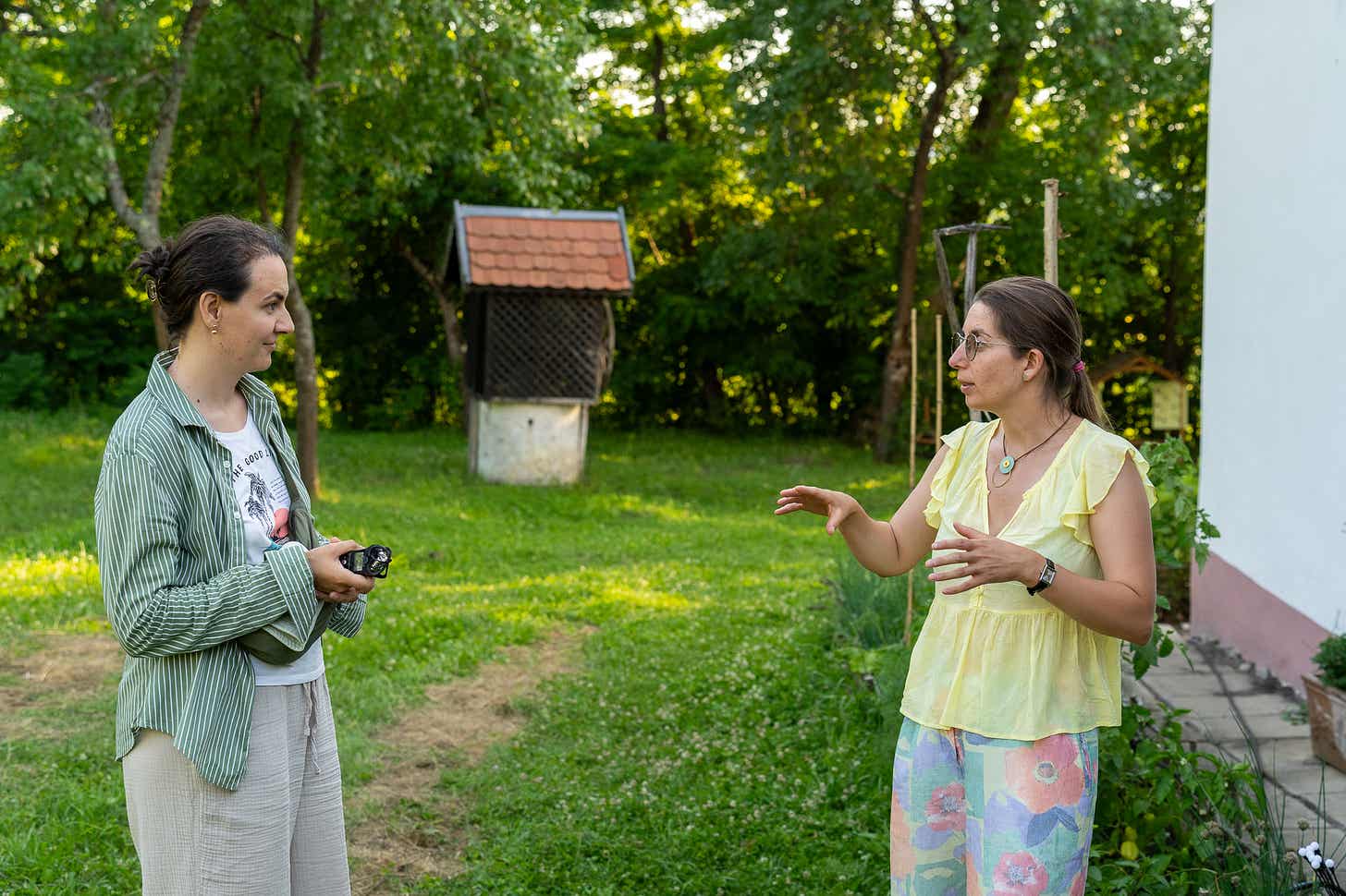 Două femei stau de vorbă pe iarbă, în fața unei case albe; una dintre ele ține un aparat foto