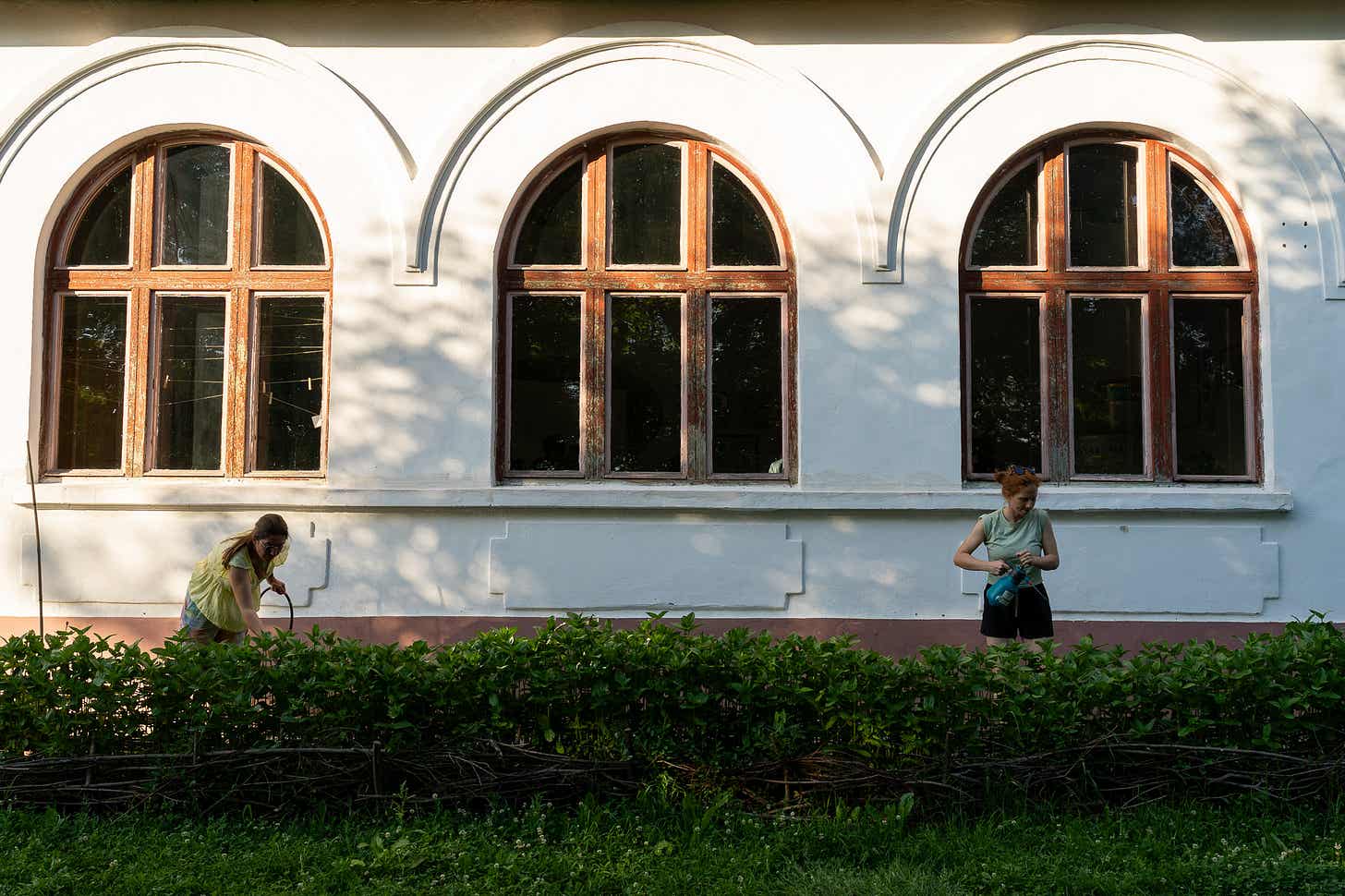 Fațada unei clădiri cu trei ferestre arcuite; două persoane lucrează la plantele din fața zidului