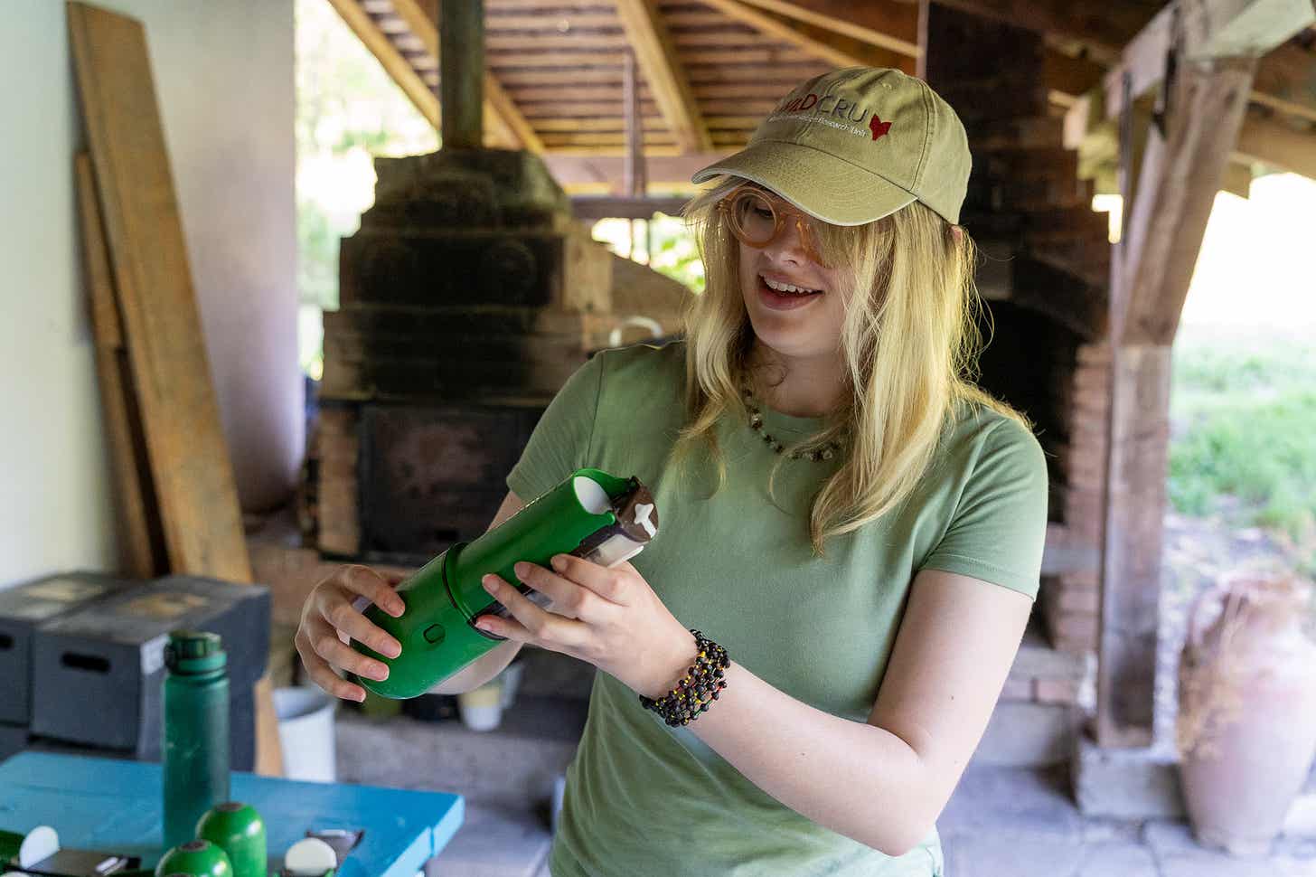 Tânără cu șapcă ține în mâini o capcană verde pentru mamifere mici, sub un șopron