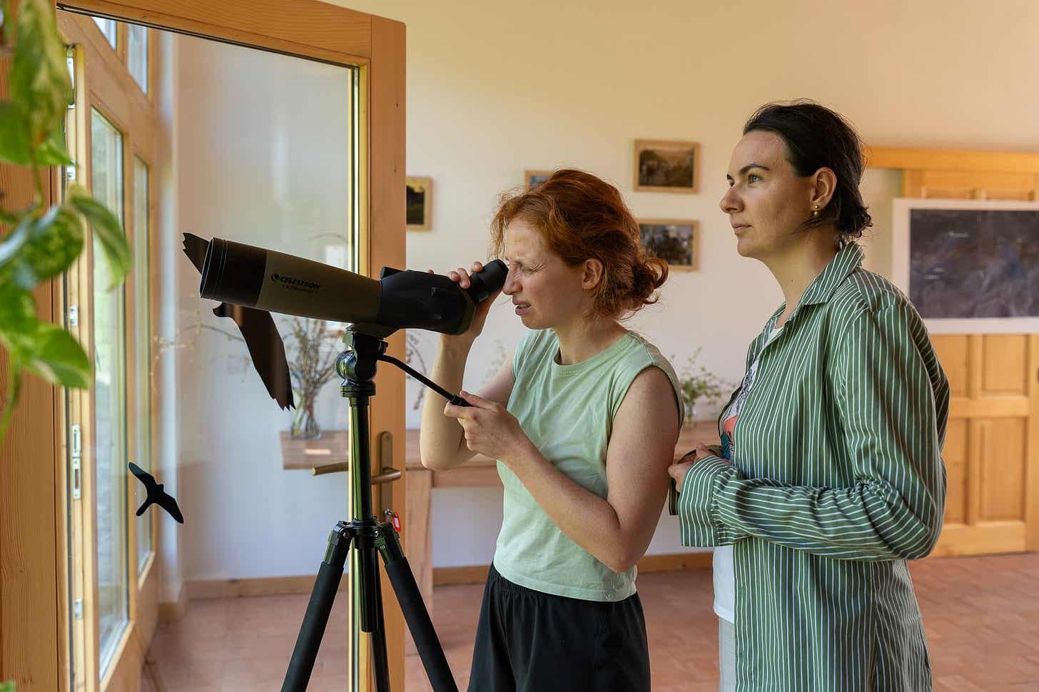 O tânără privește printr-o lunetă montată pe trepied, lângă fereastră, iar altă femeie stă alături